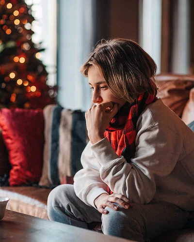 Eine Jugendliche sitzt traurig auf einem Sofa im Wohnzimmer. Sie stützt ihr Kinn auf die linke Hand. Im Hintergrund steht ein erleuchteter Weihnachtsbaum.