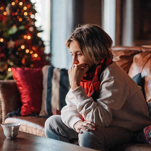 Eine Jugendliche sitzt traurig auf einem Sofa im Wohnzimmer. Sie stützt ihr Kinn auf die linke Hand. Im Hintergrund steht ein erleuchteter Weihnachtsbaum.