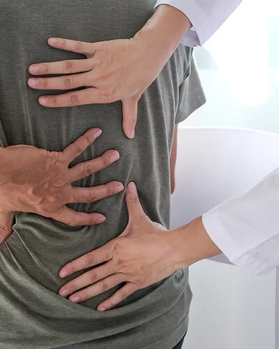 Ein Mann im T-Shirt sitzt im Behandlungszimmer einer Arztpraxis und fasst sich mit der linken Hand an den Rücken. Eine Ärztin tastet mit beiden Händen seinen Rücken ab.