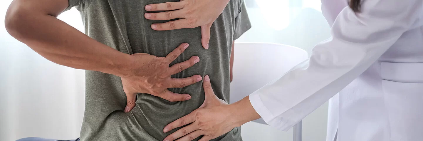 Ein Mann im T-Shirt sitzt im Behandlungszimmer einer Arztpraxis und fasst sich mit der linken Hand an den Rücken. Eine Ärztin tastet mit beiden Händen seinen Rücken ab.