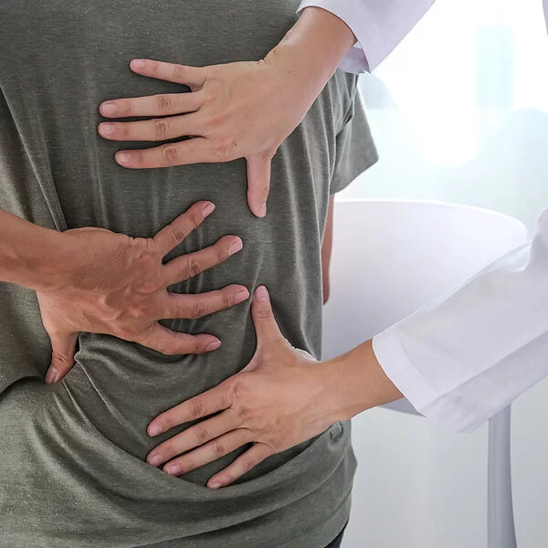 Ein Mann im T-Shirt sitzt im Behandlungszimmer einer Arztpraxis und fasst sich mit der linken Hand an den Rücken. Eine Ärztin tastet mit beiden Händen seinen Rücken ab.