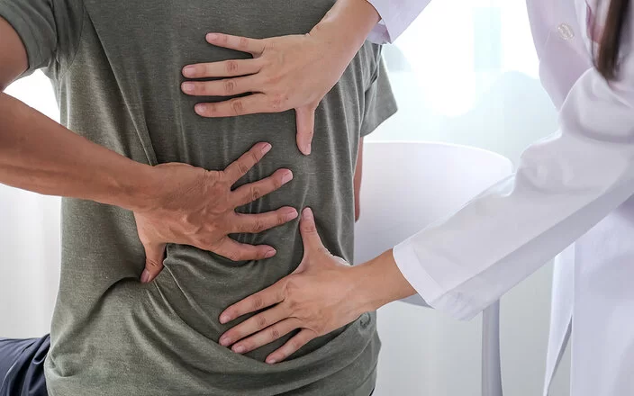 Ein Mann im T-Shirt sitzt im Behandlungszimmer einer Arztpraxis und fasst sich mit der linken Hand an den Rücken. Eine Ärztin tastet mit beiden Händen seinen Rücken ab.