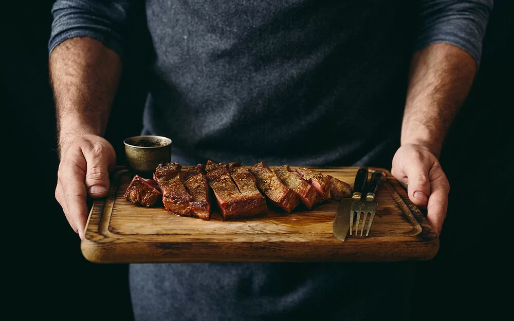 Ein Mann hält ein Schneidebrett mit einem gegrillten und in Scheiben geschnittenen Rindersteak.