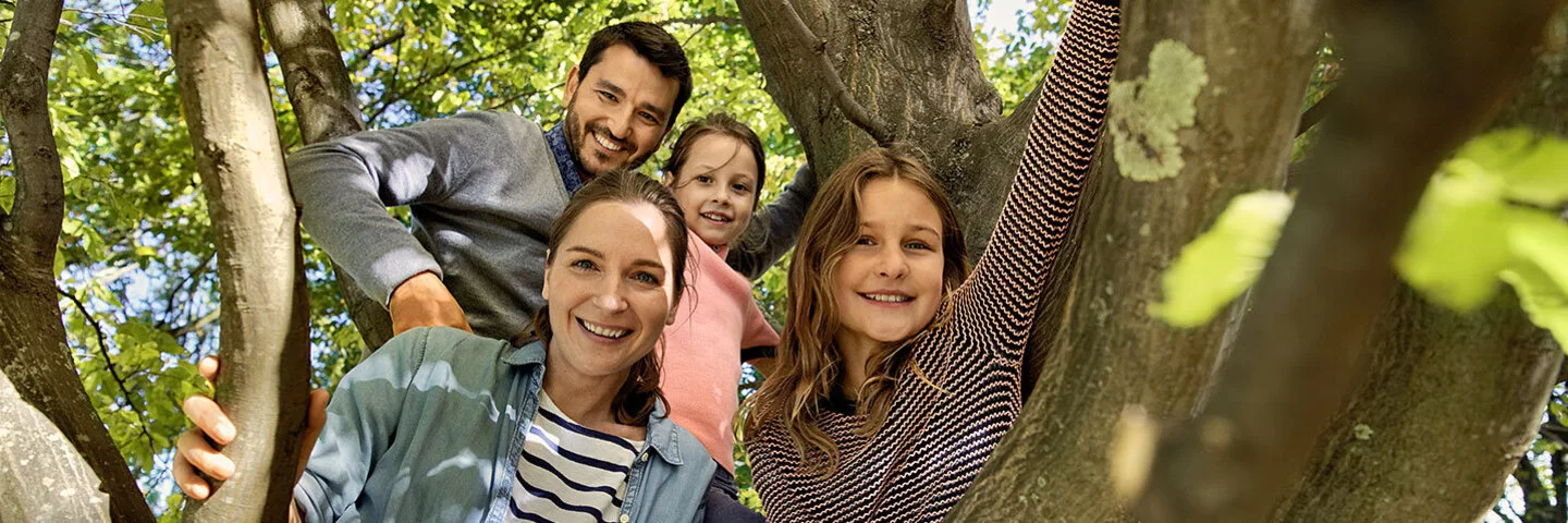 Eine Familie mit zwei Kindern ist in einen Baum geklettert und lächelt nun herunter in die Kamera. Der dunkelhaarige Vater trägt ein graues Hemd und einen Vollbart. Die Mutter trägt ein schwarz-weiß gestreiftes T-Shirt und darüber ein offenes Jeanshemd, ihre dunkelbraunen Haare trägt sie in einem Pferdeschwanz. Die beiden Töchter sind ebenfalls dunkelhaarig. Eine trägt ein rosa T-Shirt, die andere ein rosa-schwarz gestreiftes Langarm-Shirt, eine dunkelrote Shorts und eine rosa Strumpfhose. 