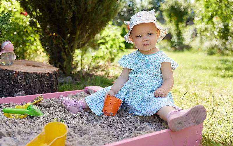 Eine kleines Mädchen in einem leichten Sommerkleid mit blauen Punkten und einem Sonnenhut aus weißem Stoff sitzt in einer rosafarbenen Plastiksandkiste mit bunten Sandförmchen und schaut in die Kamera.