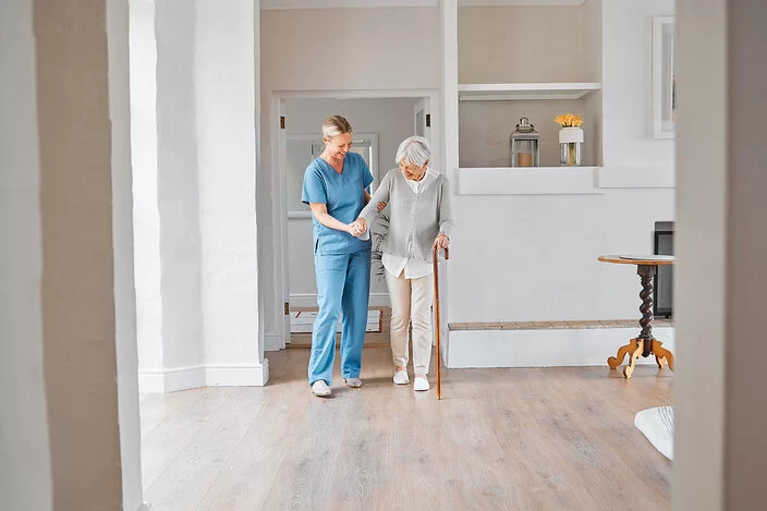 Eine Pflegerin in blauer Arbeitskleidung hilft einer Seniorin mit Gehstock beim Laufen. Sie befinden sich in einem großen, hellen Raum.