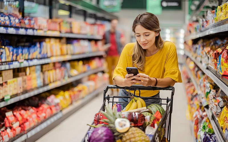 Eine junge Frau in einem gelben T-Shirt und langen dunkelblonden Haaren lehnt sich im Gang eines Supermarktes mit den Ellenbogen auf den Griff ihres gefüllten Einkaufswagens und schaut auf ihr Smartphone.