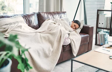 Eine junge Frau liegt mit geschlossenen Augen, angezogenen Beinen und in eine Decke gewickelt auf einer Couch und sieht leidend aus.