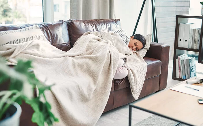 Eine junge Frau liegt mit geschlossenen Augen, angezogenen Beinen und in eine Decke gewickelt auf einer Couch und sieht leidend aus.