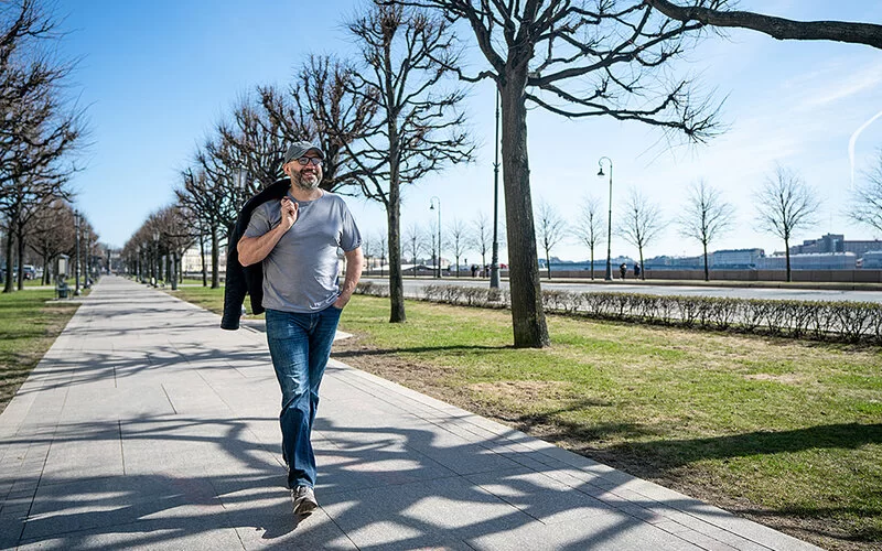 Ein vergnügter Mann mittleren Alters geht an einem Flussufer auf einem Weg entlang. Bäume, die gerade anfangen auszuschlagen, stehen links und rechts des Weges. Der Mann trägt eine Brille, einen dunklen, leicht angegrauten Vollbart und ein Basecap. Er hat die linke Hand in die Hosentasche seiner Jeans gesteckt und sich eine Jacke über die rechte Schulter geworfen und den Zeigefinger seiner rechten Hand in den Kragen gehakt, um sie festzuhalten.