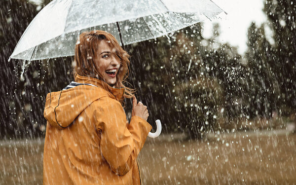 Alt-Text	Eine fröhliche Frau, die bei strömendem Regen mit einem durchsichtigen Regenschirm spazieren geht. Die Frau hat halblange rötlich-hellbraune Haare und trägt einen dunkelgelben Regenmantel. Sie hat den Betrachtenden den Rücken zugewandt und lächelt mit offenem Mund über die Schulter.