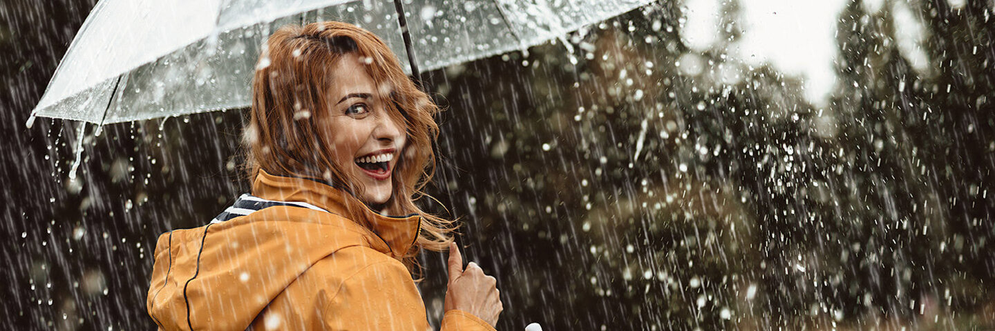 Alt-Text	Eine fröhliche Frau, die bei strömendem Regen mit einem durchsichtigen Regenschirm spazieren geht. Die Frau hat halblange rötlich-hellbraune Haare und trägt einen dunkelgelben Regenmantel. Sie hat den Betrachtenden den Rücken zugewandt und lächelt mit offenem Mund über die Schulter.