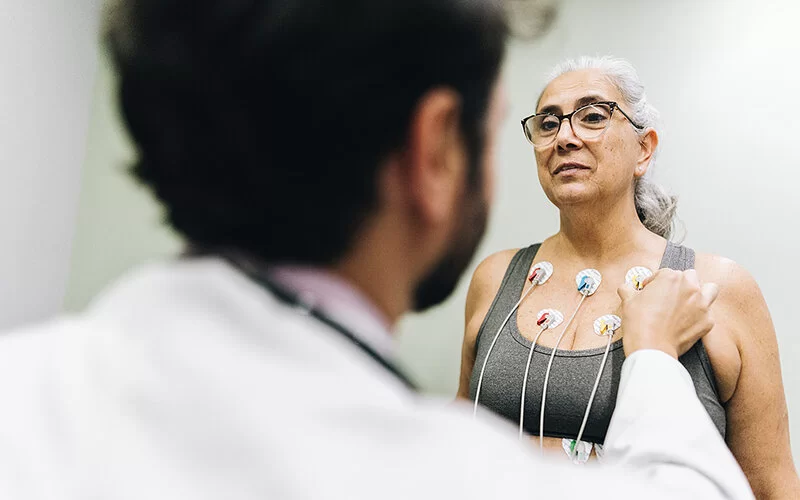 Eine Frau mittleren Alters beim Arztbesuch. Man sieht nur ihren Oberkörper, sie trägt ein graues Bustier. Ein Arzt, den man nur von hinten sieht, klebt ihr in Vorbereitung für ein EKG mehrere Kontakte oberhalb der Brust auf die Haut.