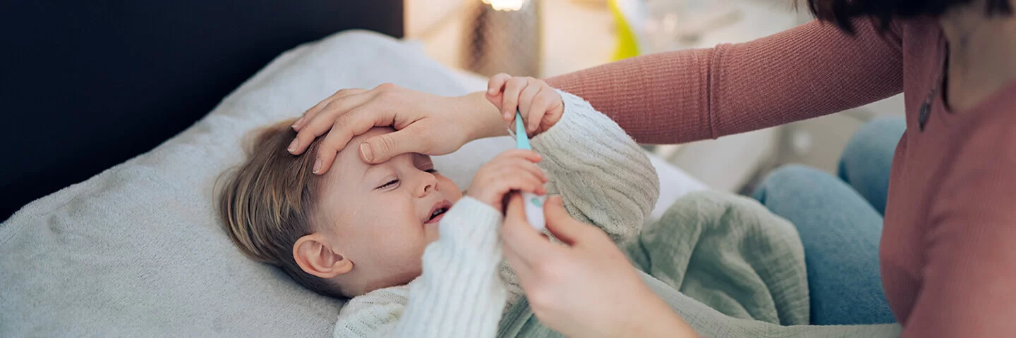 Eine Mutter nimmt die Temperatur ihres kleinen Kindes, das im Bett liegt und krank wirkt. Sie legt ihre Hand auf seine Stirn.