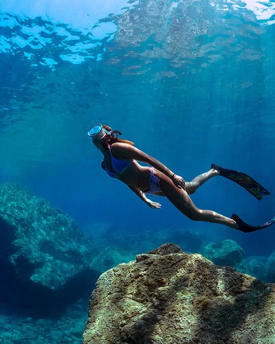 Eine Frau mit Taucherbrille und Flossen schwimmt unter Wasser über große Felsbrocken hinweg.