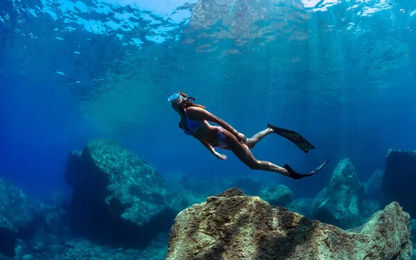 Eine Frau mit Taucherbrille und Flossen schwimmt unter Wasser über große Felsbrocken hinweg.