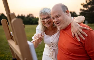 Eine ältere Frau und ein etwa 40 bis 55 Jahre alter Mann mit Down-Syndrom stehen in der Abendsonne auf einer Wiese vor einer Staffelei. Die Frau führt mit der rechten Hand einen Pinsel über die Leinwand und hat ihren linken Arm über die Schultern des Mannes gelegt. Beide lächeln.