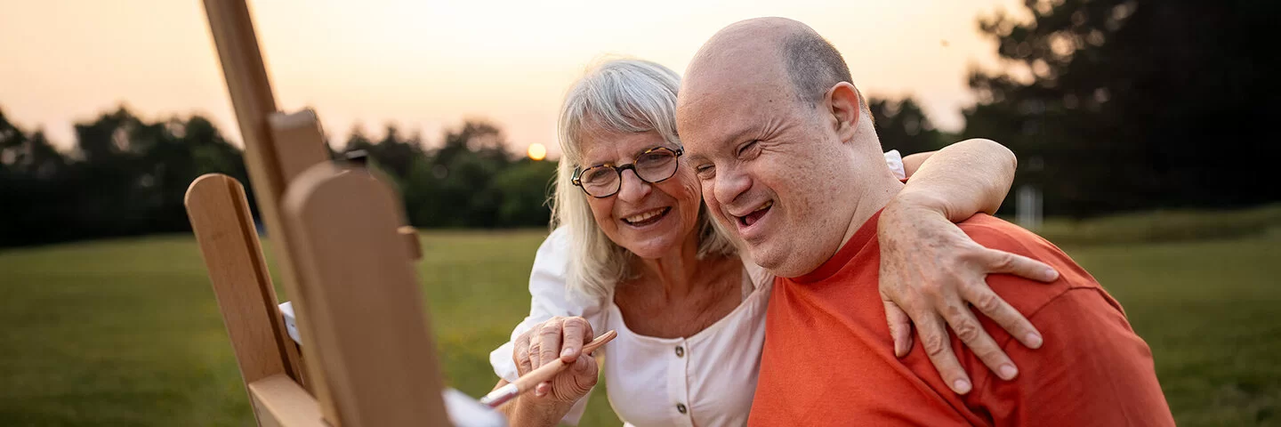 Eine ältere Frau und ein etwa 40 bis 55 Jahre alter Mann mit Down-Syndrom stehen in der Abendsonne auf einer Wiese vor einer Staffelei. Die Frau führt mit der rechten Hand einen Pinsel über die Leinwand und hat ihren linken Arm über die Schultern des Mannes gelegt. Beide lächeln.