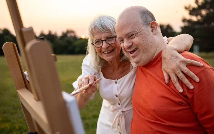 Eine ältere Frau und ein etwa 40 bis 55 Jahre alter Mann mit Down-Syndrom stehen in der Abendsonne auf einer Wiese vor einer Staffelei. Die Frau führt mit der rechten Hand einen Pinsel über die Leinwand und hat ihren linken Arm über die Schultern des Mannes gelegt. Beide lächeln.