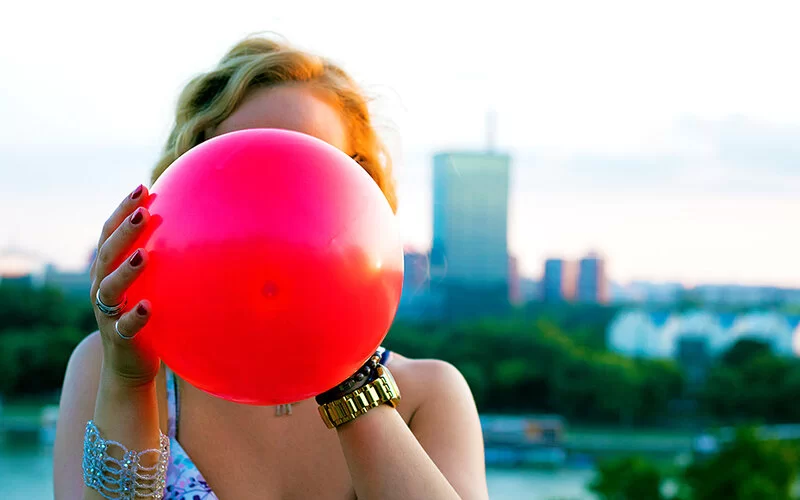 Sommer in der Großstadt. Nahaufnahme einer Frau, die einen roten Luftballon aufbläst. Das Gesicht der Frau ist hinter dem Luftballon verborgen.