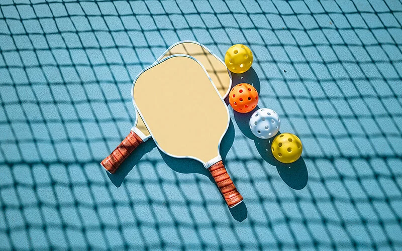 Zwei Pickleball-Schläger und vier bunte Bälle liegen auf dem Boden.