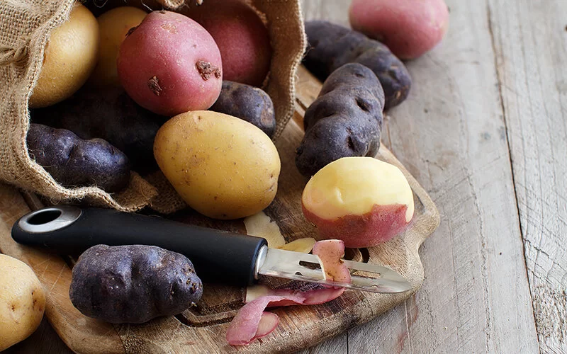 Auf einem Brett liegen bunte Kartoffeln und ein Schälmesser.