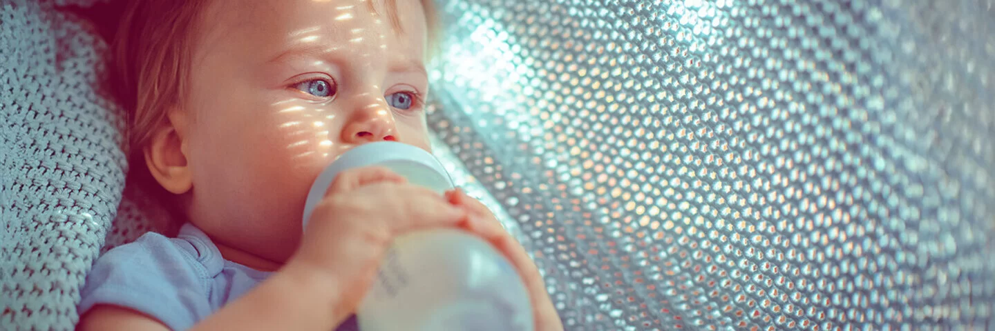 Nahaufnahme von einem Kleinkind, das aus einer Flasche trinkt und in einer hellblauen gestrickten Decke liegt.