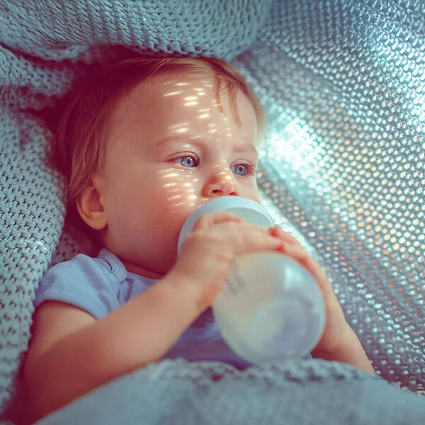 Nahaufnahme von einem Kleinkind, das aus einer Flasche trinkt und in einer hellblauen gestrickten Decke liegt.