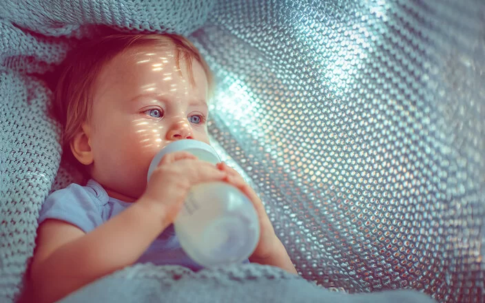 Nahaufnahme von einem Kleinkind, das aus einer Flasche trinkt und in einer hellblauen gestrickten Decke liegt.