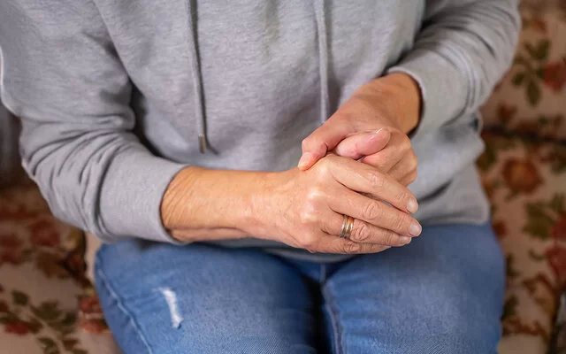 Eine Frau in sitzender Position umschließt mit einer Hand den Daumen der anderen Hand.