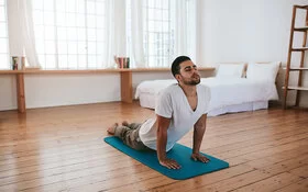 Ein junger Mann macht zuhause Yoga-Übungen. Mit den Händen stützt er sich auf einer Gymnastikmatte ab und hebt den Oberkörper. Der Kopf ist leicht nach hinten geneigt, die Augen sind geschlossen, Seine Beine sind auf der Matte abgelegt. 