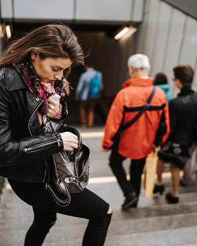 Eine junge Frau durchsucht auf den Treppen, die zu einer U-Bahn-Station hinabführen, ihre Handtasche. Die Frau hat offene, lange dunkelbraune Haare und trägt eine schwarze Lederjacke und enge, schwarze Hosen mit einem Loch im rechten Knie.