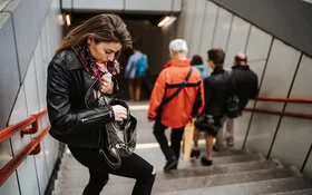 Eine junge Frau durchsucht auf den Treppen, die zu einer U-Bahn-Station hinabführen, ihre Handtasche. Die Frau hat offene, lange dunkelbraune Haare und trägt eine schwarze Lederjacke und enge, schwarze Hosen mit einem Loch im rechten Knie.