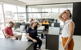 Eine Gruppe von Büroangestellten sitzt an ihren Schreibtischen und dehnt den Nacken. Eine Frau steht rechts im Bild und macht die Übung vor.