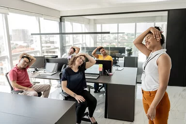 Eine Gruppe von Büroangestellten sitzt an ihren Schreibtischen und dehnt den Nacken. Eine Frau steht rechts im Bild und macht die Übung vor.