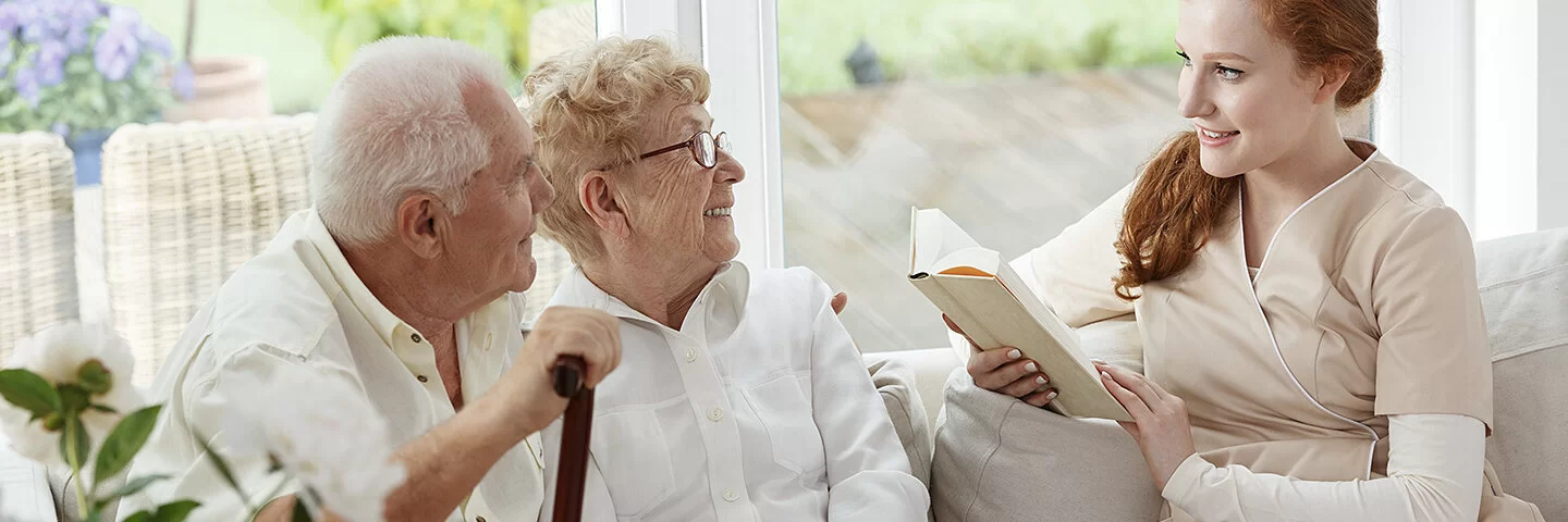 Eine junge Frau liest einem Paar Senioren aus einem Buch vor.