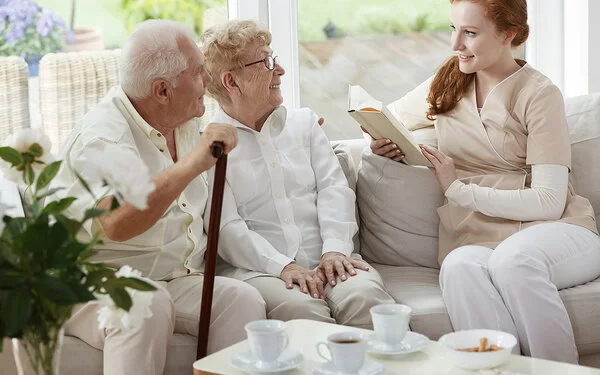 Eine junge Frau liest einem Paar Senioren aus einem Buch vor.