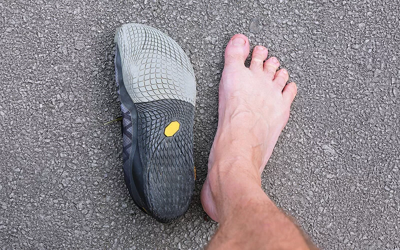 Nahaufnahme auf einen Fuß, der auf Asphalt steht, daneben liegt ein Barfußschuh.