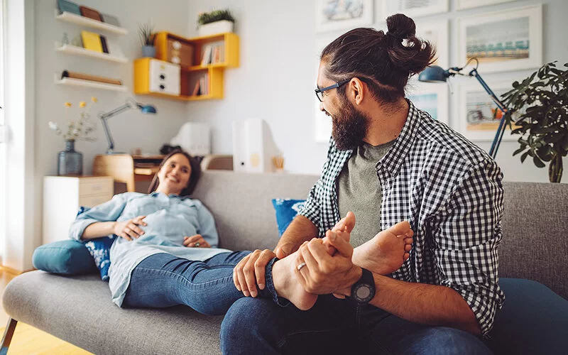 Eine schwangere Frau und ihr Partner sitzen auf dem Sofa. Sie hat die Beine hochgelegt. Er umfasst ihren rechten Unterschenkel und den rechten Fuß.