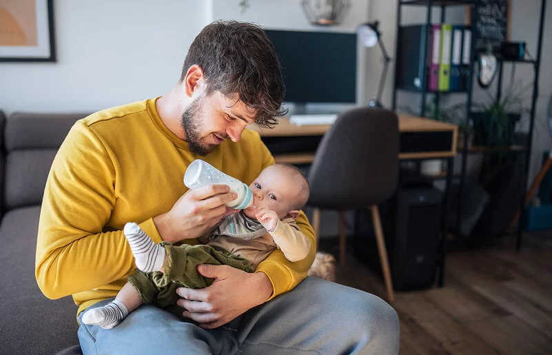 Ein junger Mann mit braunen Haaren und einem kurzen Bart sitzt in einem Wohnzimmer auf der Sofakante und füttert ein Baby mit einer Flasche. Der Mann trägt einen geben Pullover und schaut auf das Kind. Das Kind trägt eine grüne Hose und schaut in die Kamera. Im Hintergrund ist ein Schreibtisch zu sehen.