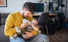 Ein junger Mann mit braunen Haaren und einem kurzen Bart sitzt in einem Wohnzimmer auf der Sofakante und füttert ein Baby mit einer Flasche. Der Mann trägt einen geben Pullover und schaut auf das Kind. Das Kind trägt eine grüne Hose und schaut in die Kamera. Im Hintergrund ist ein Schreibtisch zu sehen.