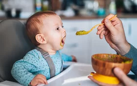 Ein Baby sitzt mit weit geöffnetem Mund in einem Hochstuhl und wird von einem Mann, der nicht im Bild zu sehen ist, mit einem grünen Gemüsebrei gefüttert.