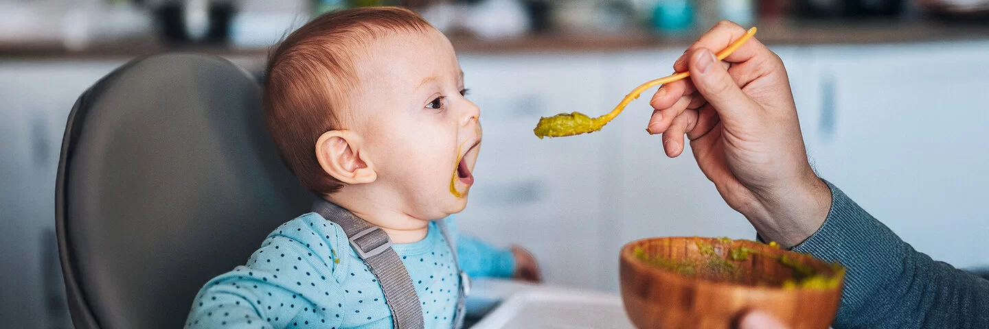 Ein Baby sitzt mit weit geöffnetem Mund in einem Hochstuhl und wird von einem Mann, der nicht im Bild zu sehen ist, mit einem grünen Gemüsebrei gefüttert.