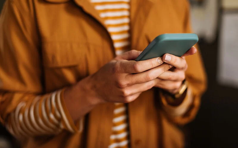 Nahaufnahme einer Frau in einem gestreiften Oberteil mit einer Jacke darüber, die ein Smartphone in der Hand hält.