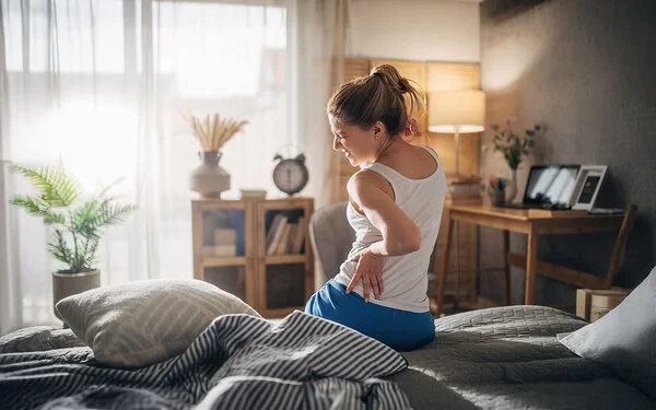 Eine Frau sitzt auf dem Bett und hält sich mit schmerzverzerrtem Gesicht den Rücken.