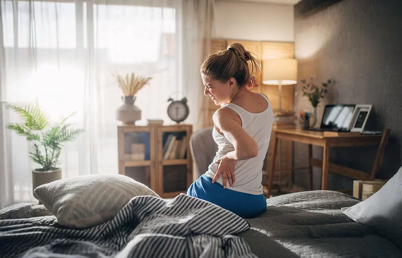 Eine Frau sitzt auf dem Bett und hält sich mit schmerzverzerrtem Gesicht den Rücken.