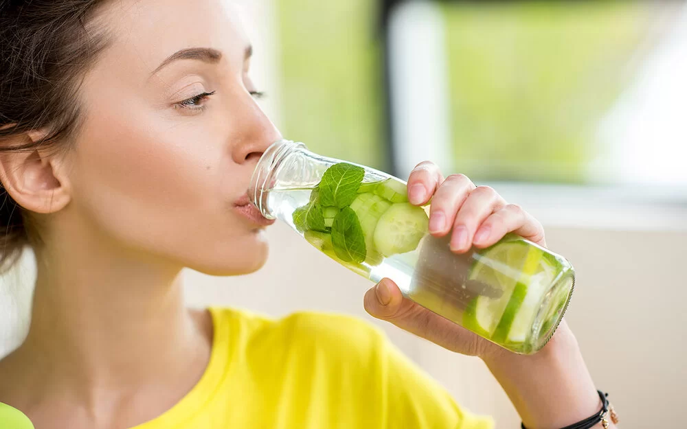 Eine junge Frau mit zurückgebundenen braunen Haaren trinkt aus einer Glasflasche Wasser, das mit Minzblättern sowie Gurken- und Limettenscheiben aromatisiert ist.