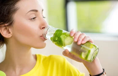 Eine junge Frau mit zurückgebundenen braunen Haaren trinkt aus einer Glasflasche Wasser, das mit Minzblättern sowie Gurken- und Limettenscheiben aromatisiert ist.