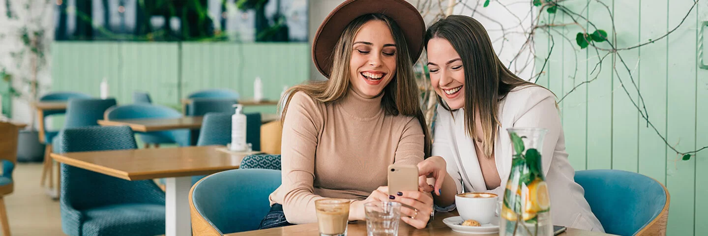 Zwei junge Frauen sitzen lachend an einem Tisch und schauen auf ein Handy.