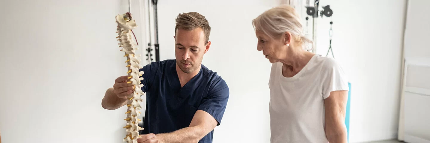 Eine ältere Frau sitzt in Sportkleidung auf einem Gymnastikball, ein Physiotherapeut erklärt ihr etwas anhand eines Skelett-Modells.